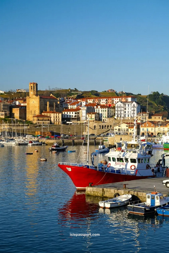 A vibrant red fishing boat moored in Getaria harbour, with a medieval hilltop village and a historic stone church tower rising behind it along the rugged Basque coast of Northern Spain.