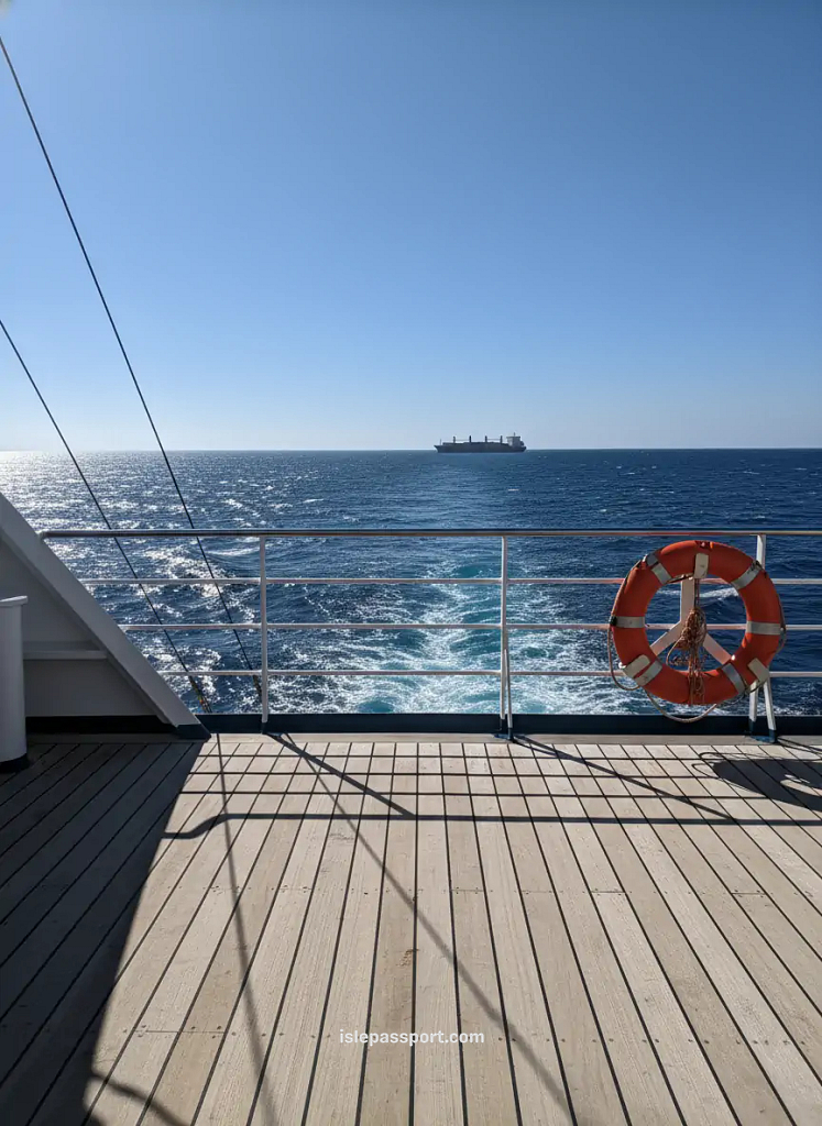 Midday ferry deck with harsh sun, bright reflections on metal railings, deep-blue waves, and distant cargo ship