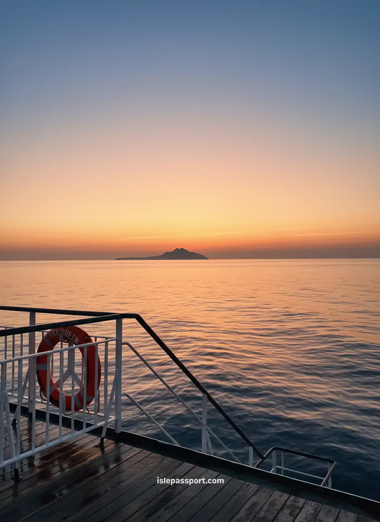 Ferry deck at sunset with golden sky gradient, calm sea reflections, and distant island silhouette