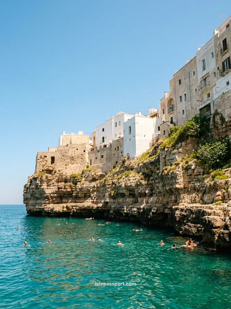 People swimming in the clear turquoise water beneath the dramatic limestone cliffs and white houses of Polignano a Mare.