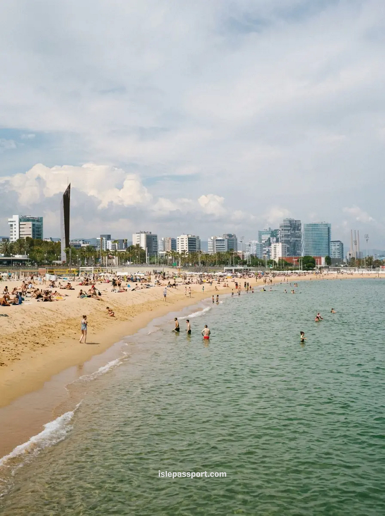 A crowded but wide sandy beach in the Poblenou district of Barcelona with modern skyscrapers in the background.