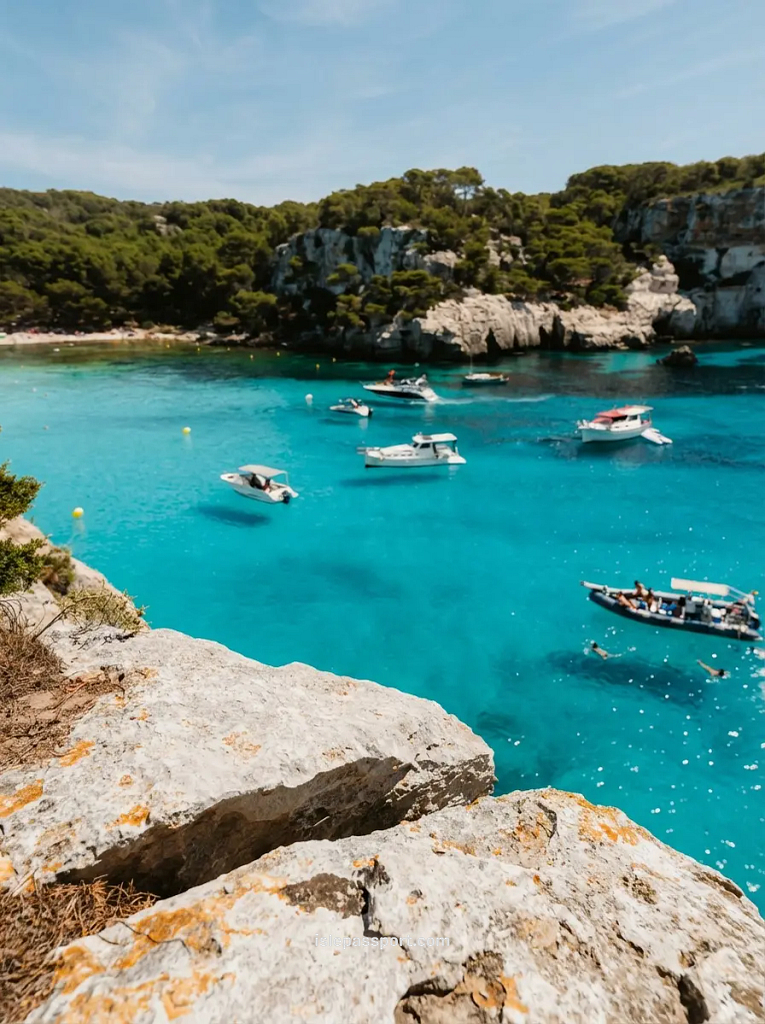 A dramatic close-up of light-colored foreground rocks overlooks a protected cove with vibrant electric turquoise water, scattered boats and yachts, and dense pine forest lining limestone cliffs at Cala Trebaluger, Menorca.