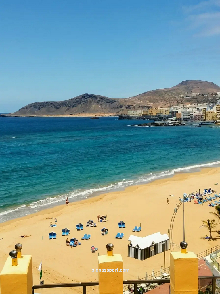 A wide, scenic shot of Playa de Las Canteras in Las Palmas, Gran Canaria, showing the bustling sandy beach with blue sun loungers, an inviting turquoise ocean bay, and the city buildings set against a backdrop of a massive volcanic mountain and a clear blue sky.