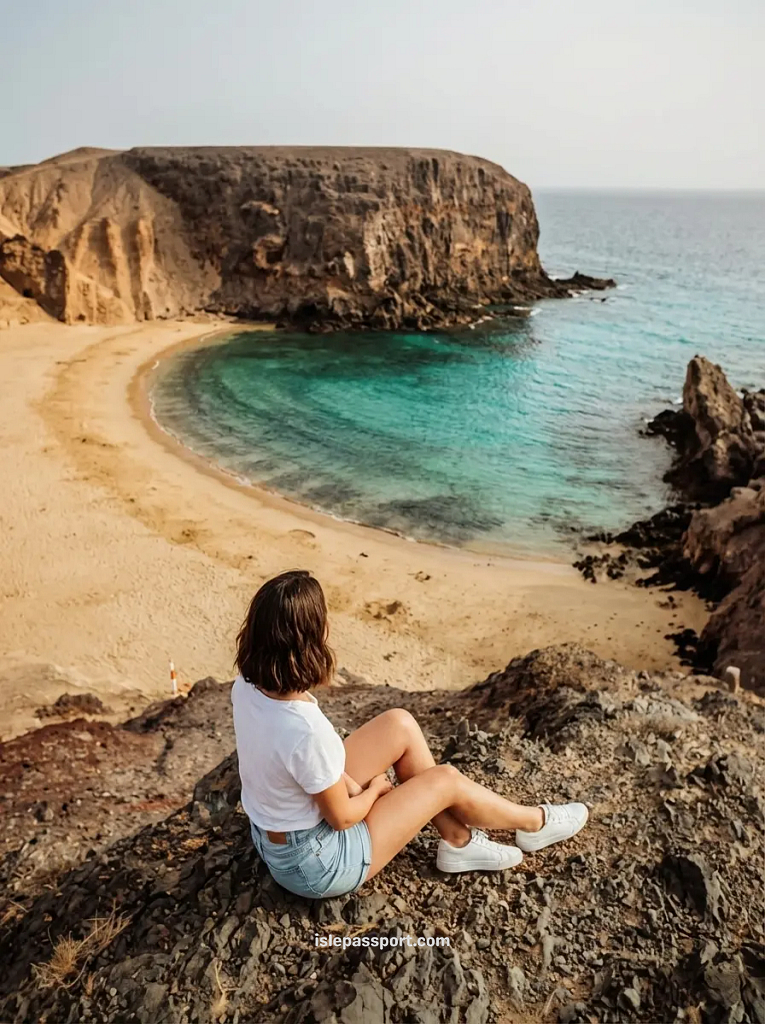 A woman sitting on a dark volcanic cliff overlooking the golden sands and turquoise cove of Playa de Papagayo in Lanzarote, Canary Islands.