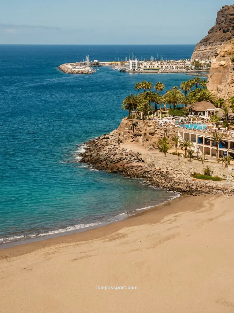 An aerial-perspective photograph looking down at the sheltered sandy bay of Playa de Mogán in Gran Canaria, featuring palm trees and blue water, leading to a marina filled with sailboats and a whitewashed resort village built against a dramatic, arid coastal cliff.