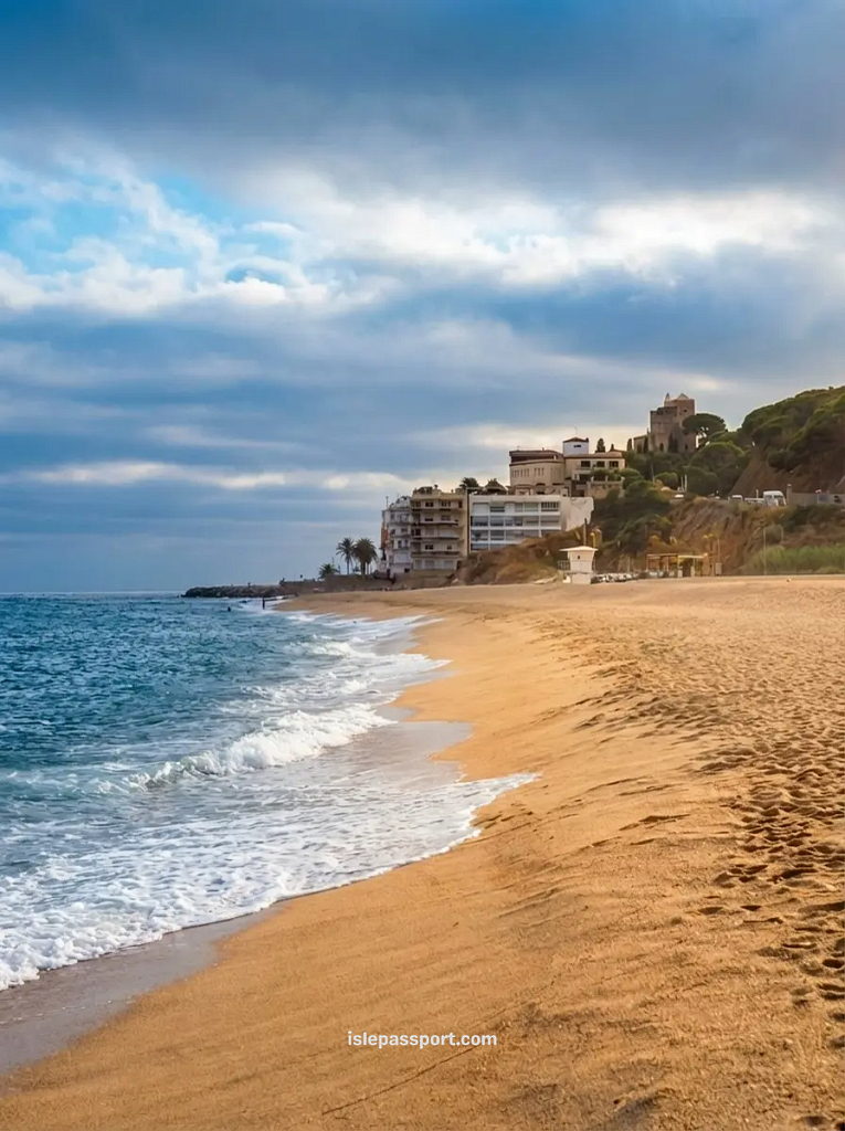 A wide view of the expansive golden sand beach in Sant Pol de Mar, leading towards a cluster of modern white buildings and an ancient watchtower on a hillside overlooking the sea.