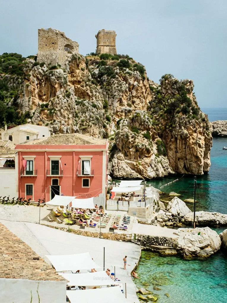 An authentic pink coastal villa in Sicily is situated directly on a rocky shoreline with clear, calm turquoise water.