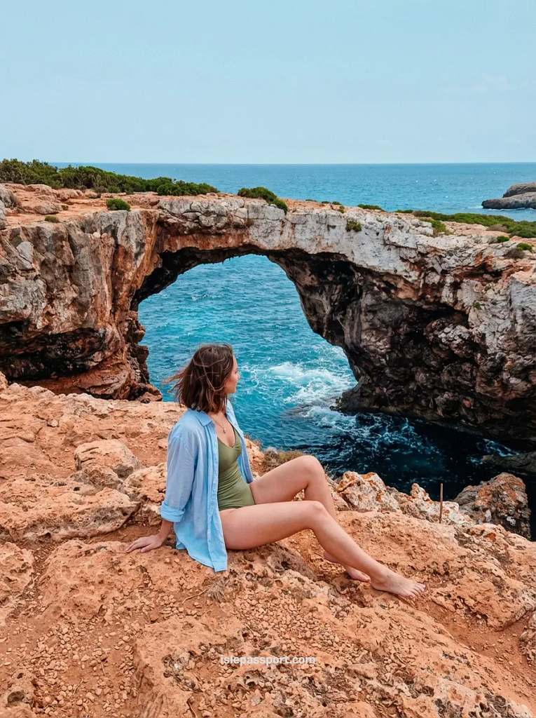 A woman sitting on a cliff overlooking the natural sea cave arch at Cala Varques, a famous spot among Mallorca hidden beaches.