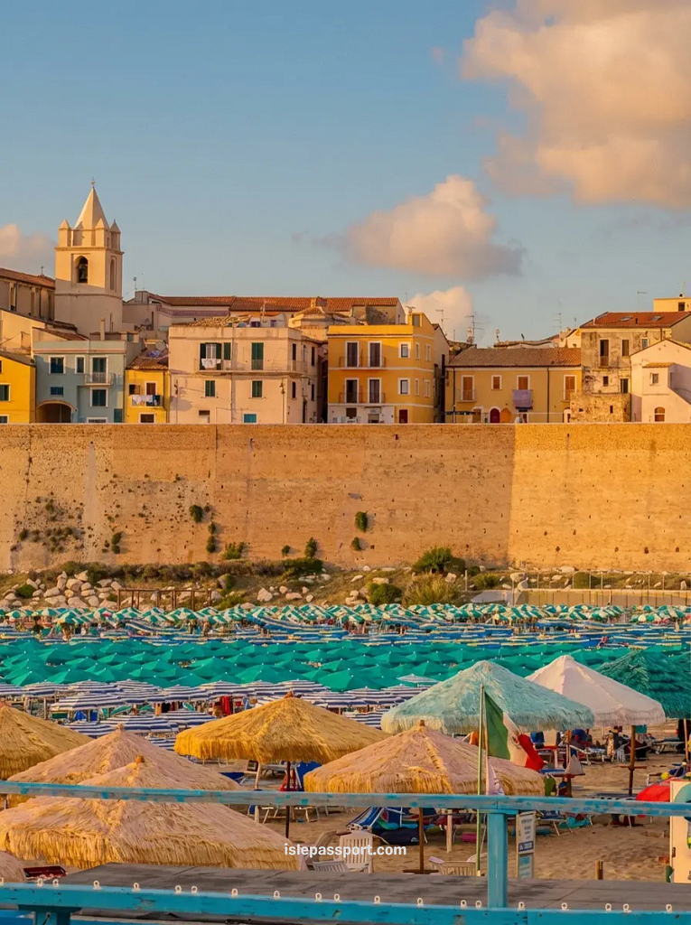 A view of the colorful historic buildings and church tower in Termoli, one of the cheap coastal towns in Italy, overlooking a beach with blue and green umbrellas.