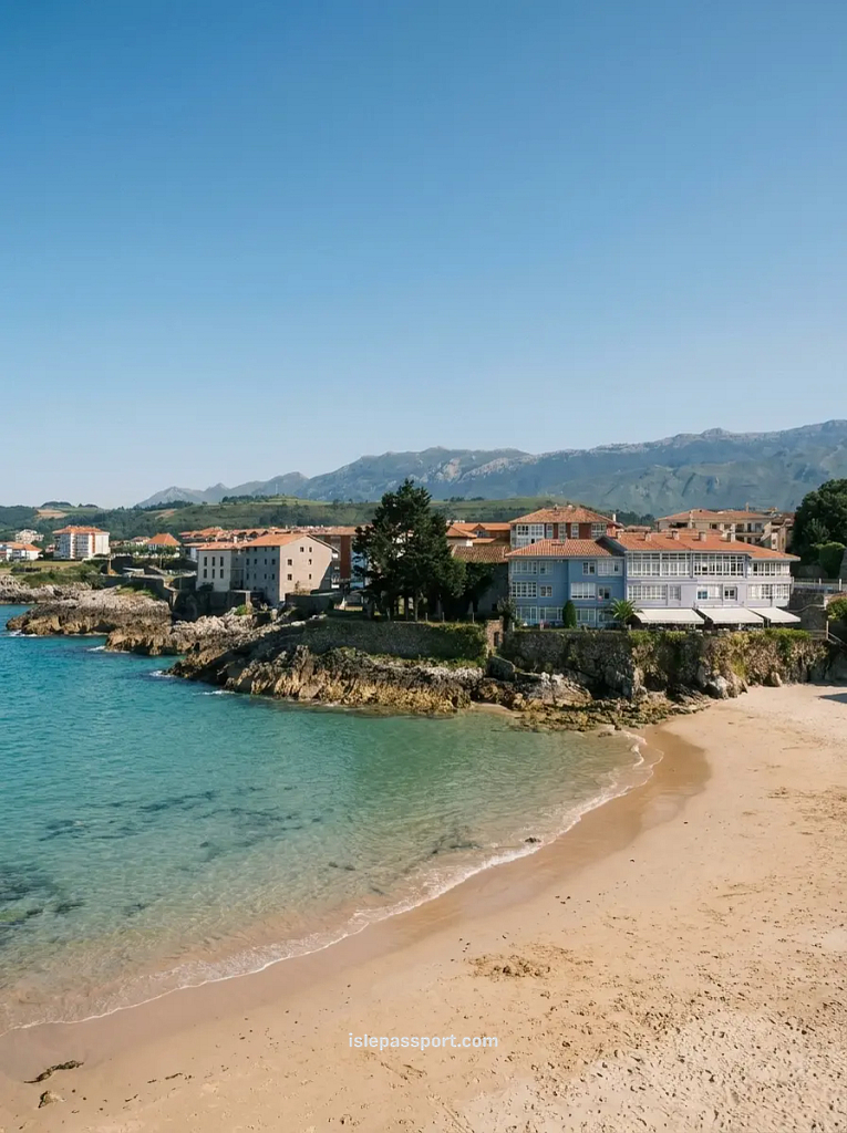 A sheltered sandy cove in Llanes with clear turquoise water, a village of white and blue buildings perched on the rocky headland, and the Picos de Europa mountains visible in the background