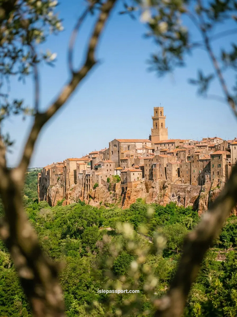 A scenic view framed by olive branches looking across a green valley at the cliffside town of Pitigliano, with stone buildings carved directly into volcanic rock.