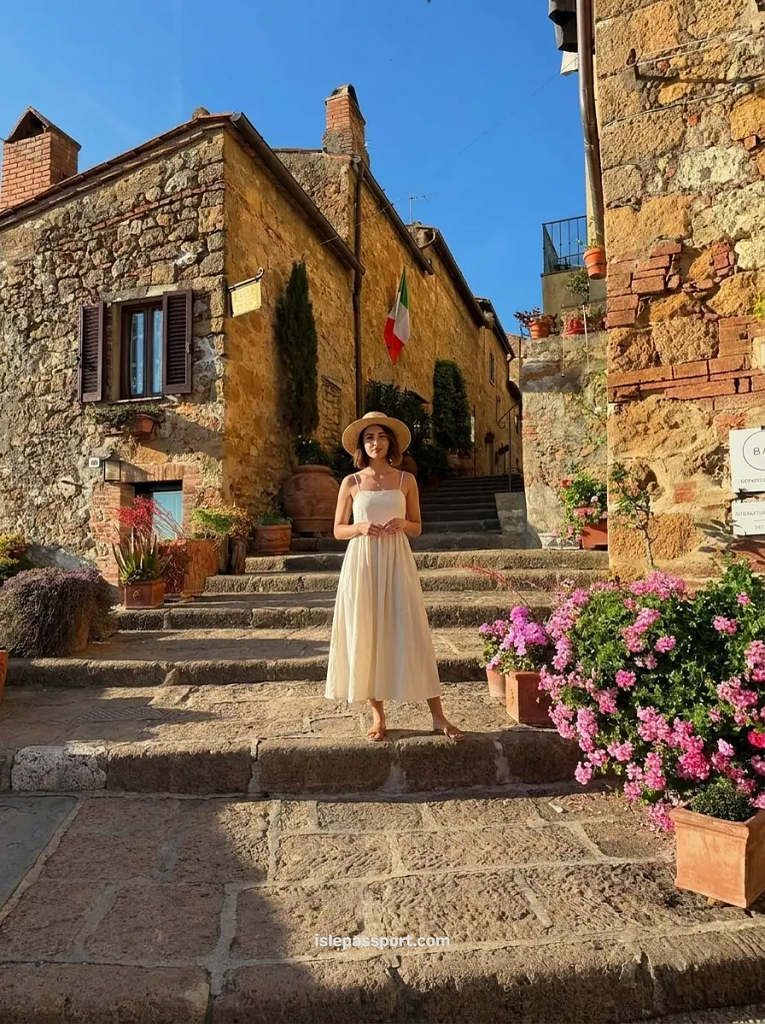 A traveler in a sun hat standing on the historic stone steps of a sun-drenched medieval village in Pienza, surrounded by vibrant pink flowers.