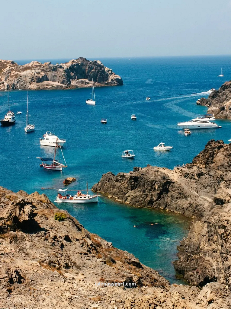 High-angle view looking down at sailboats and motorboats anchored in the deep blue and turquoise waters of Cala Jugadora in Cap de Creus Natural Park.