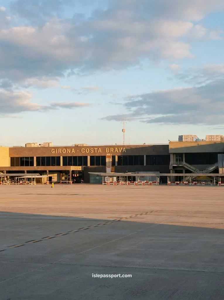 The exterior facade of the Girona-Costa Brava airport terminal under a cloudy sky during sunset golden hour.