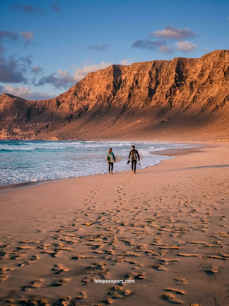Two surfers carrying boards along Famara beach at sunset, with the Famara cliffs behind, Lanzarote