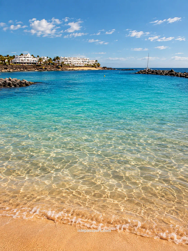 Calm turquoise water and golden sand at a sheltered beach in Costa Teguise, Lanzarote, with white apartments in the distance